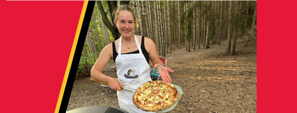 a person in an apron holds a wood fired pizza on a tra while standing among evergreen trees
