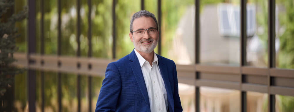 Dr. Rene Van Acker, dressed in a navy blue suit and white dress shirt, stand against a fence area on a clear sunny day