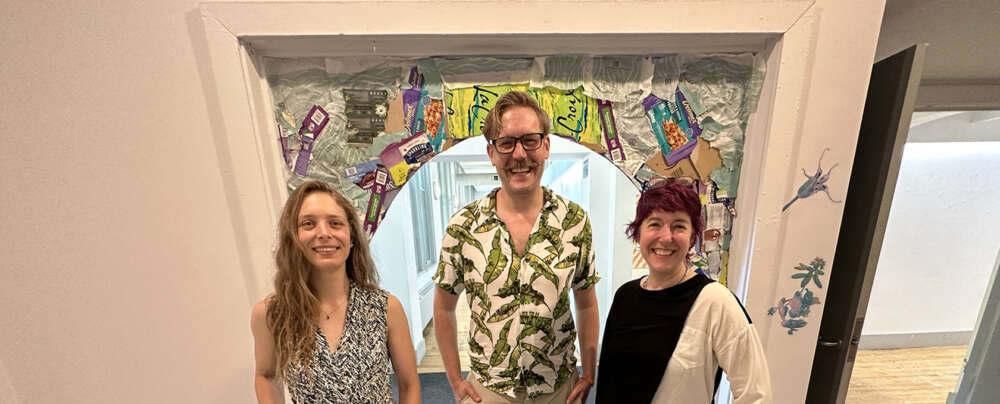 Three people in patterned clothing stand in front of an archway made from recycled materials in an art gallery at the University of Guelph.