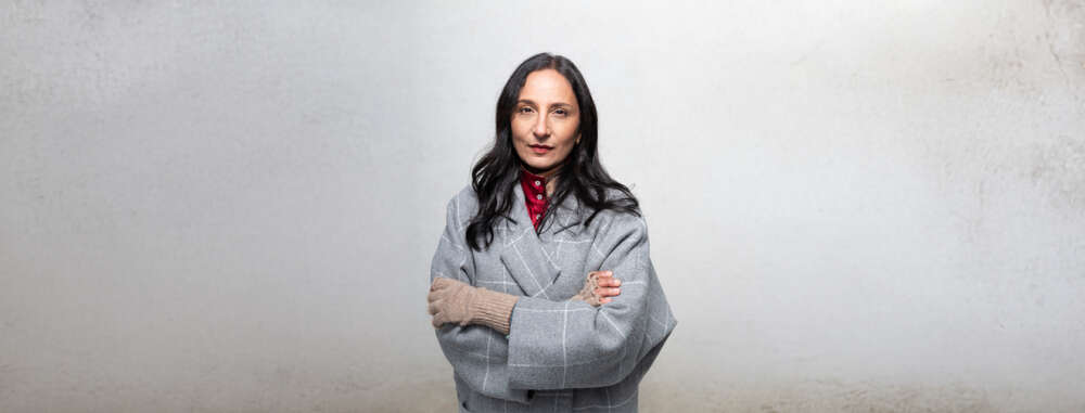 Madhur Anand, dressed in a gray coat, crosses her arms powerfully against a gray background