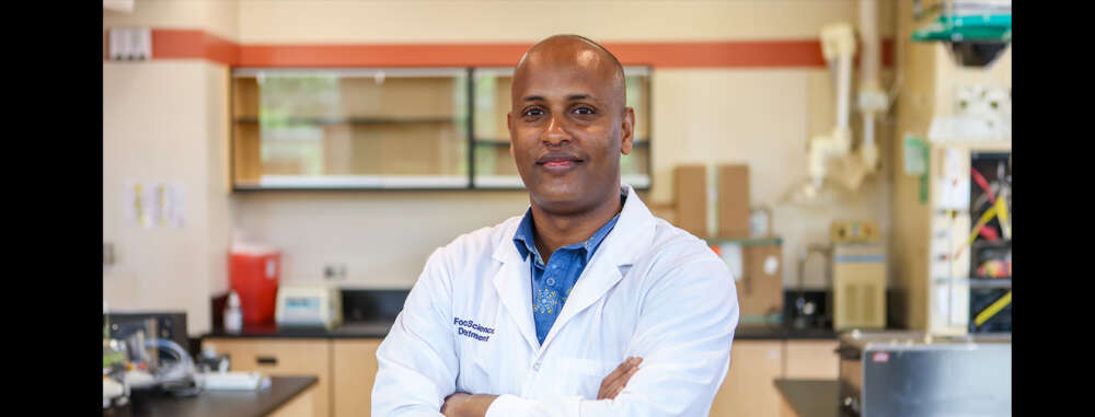 Dr.  Biniam Kebede wears a lab coat and poses for a photo in his lab
