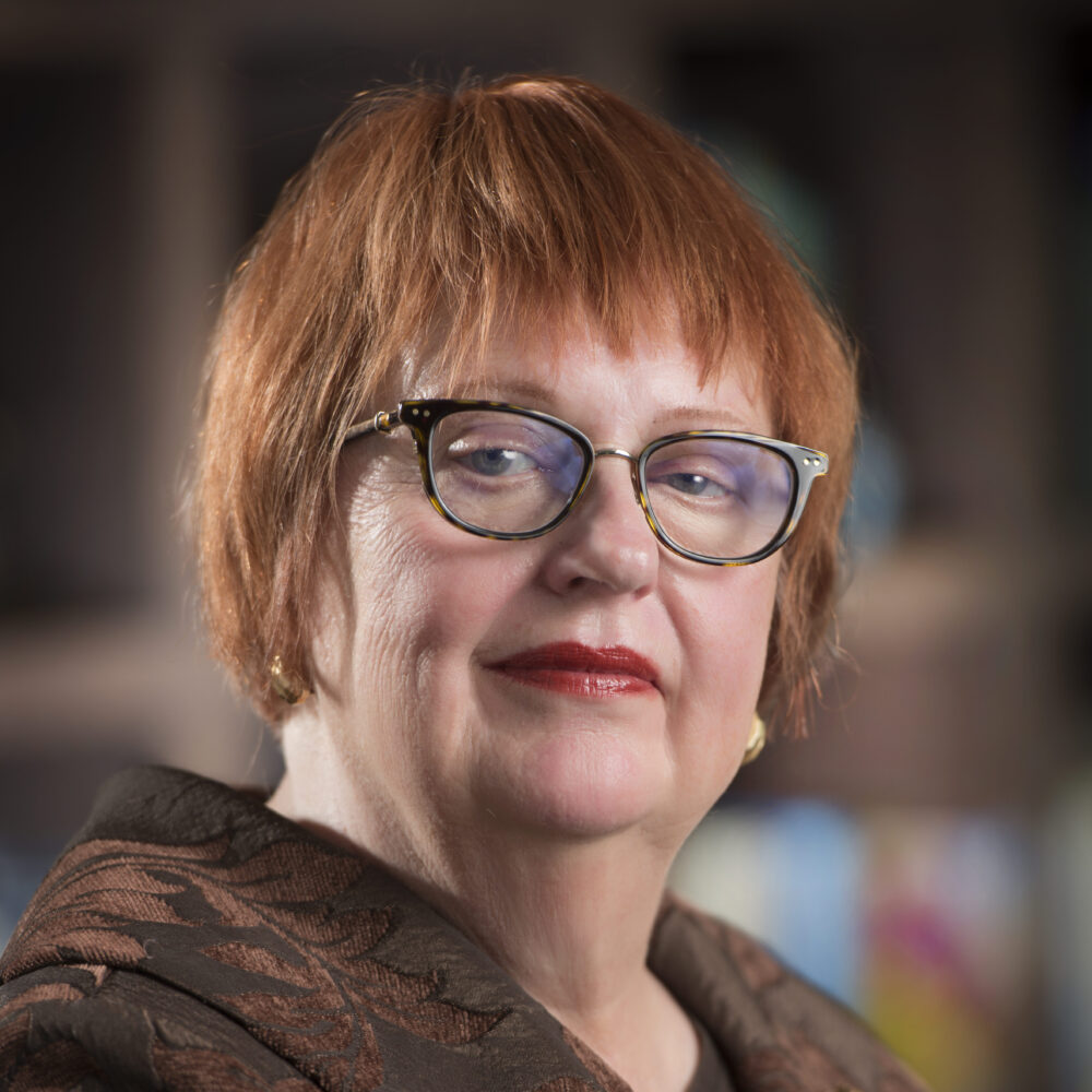a person with short red hair and glasses wearing a brown jacket poses for a headshot