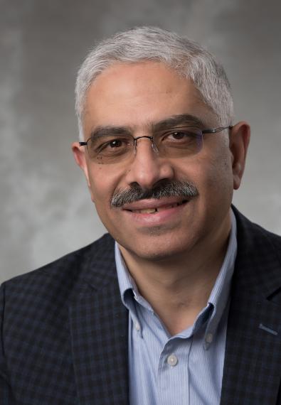 Headshot of Dr. Medhat Moussa, against a grey background, he has short white hair, glasses