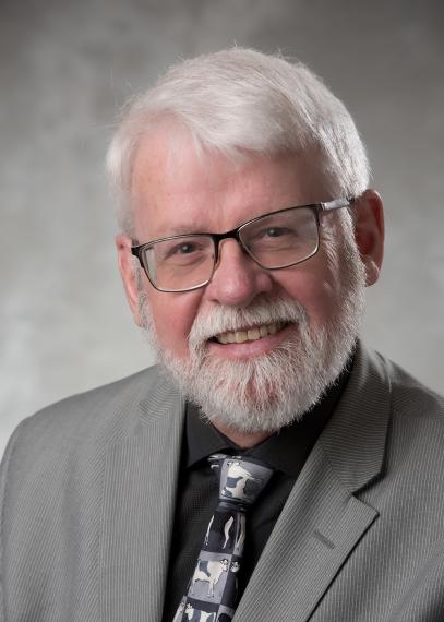 a person with grey hair and glasses in a grey blazer, black shirt and grey tie poses for a headshot