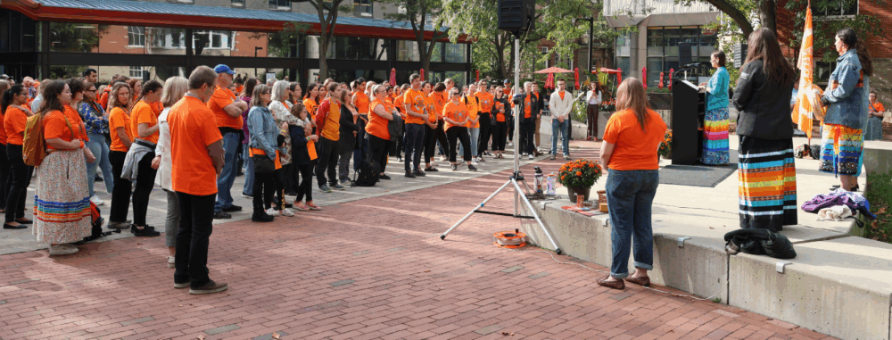 A group of people gather in Branion Plaza wearing Orange Shirts and listen to a speaker at a podium.