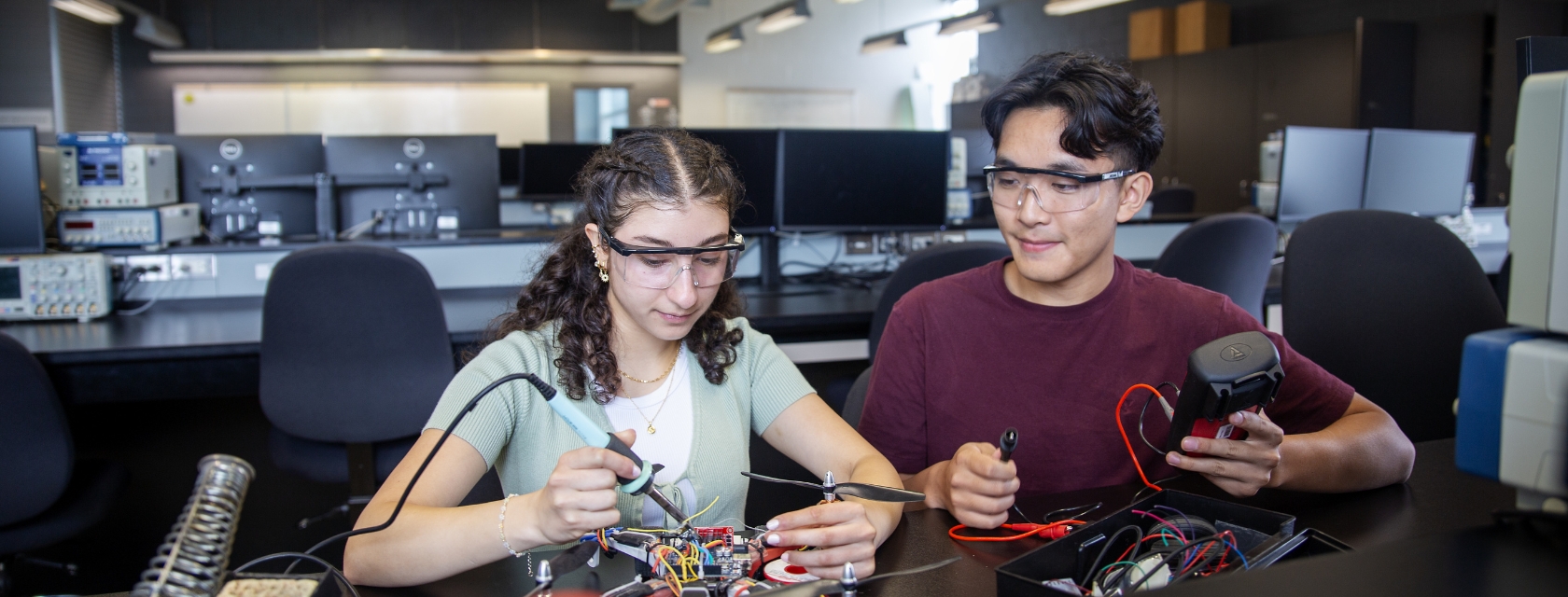 Two students wearing safety glasses work together on drone components in an engineering lab. One student uses a soldering iron while the other holds a controller with red wires. Computer monitors and testing equipment line the benches behind them.