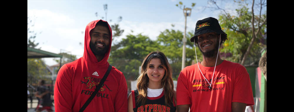 Three U of G students stand in a line wearing red and black Gryphon sports attire and smiling into the camera