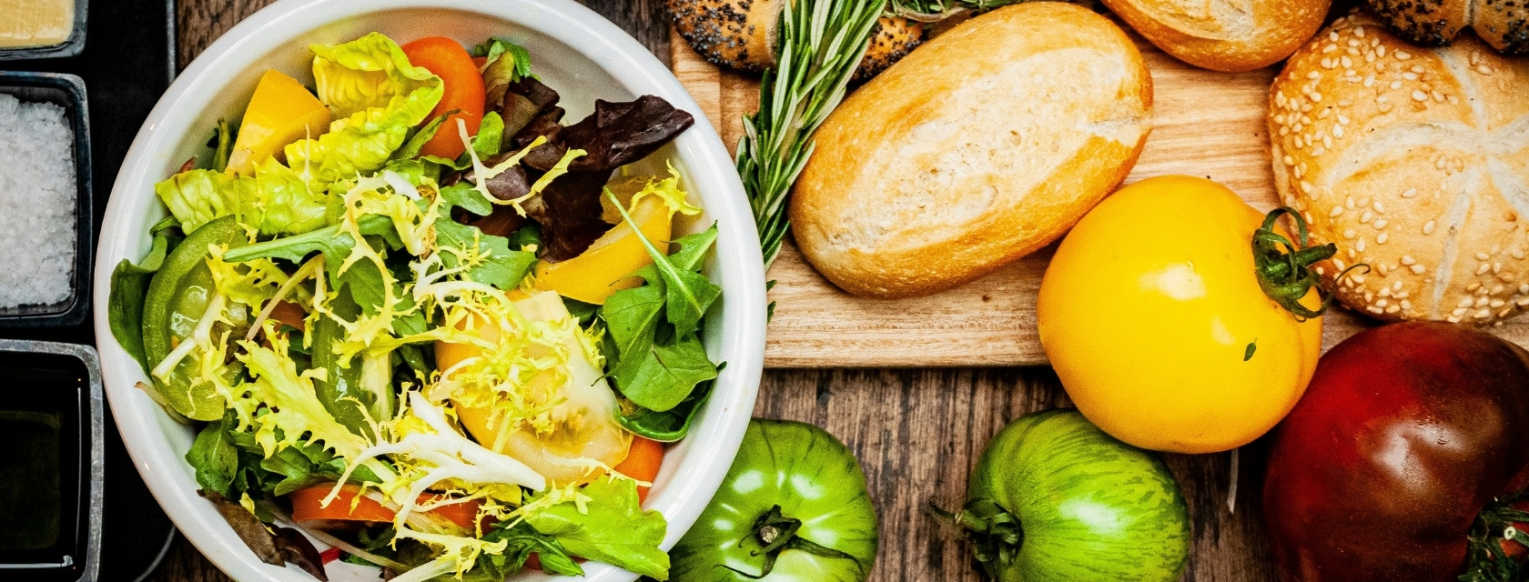 A fresh salad bowl filled with leafy greens, frisée, arugula, red leaf lettuce, and sliced heirloom tomatoes, beside a wooden board displaying a variety of rustic bread rolls, rosemary sprigs, and colourful tomatoes (Unsplash / Bern Fresen)