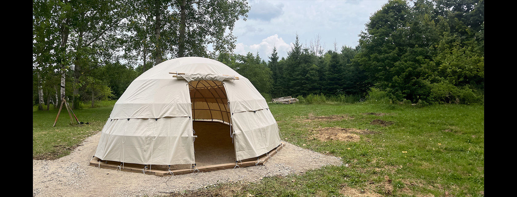 a beige canvas covered structure sits in the centre of a patch of gravel, surrounded by green grass and trees
