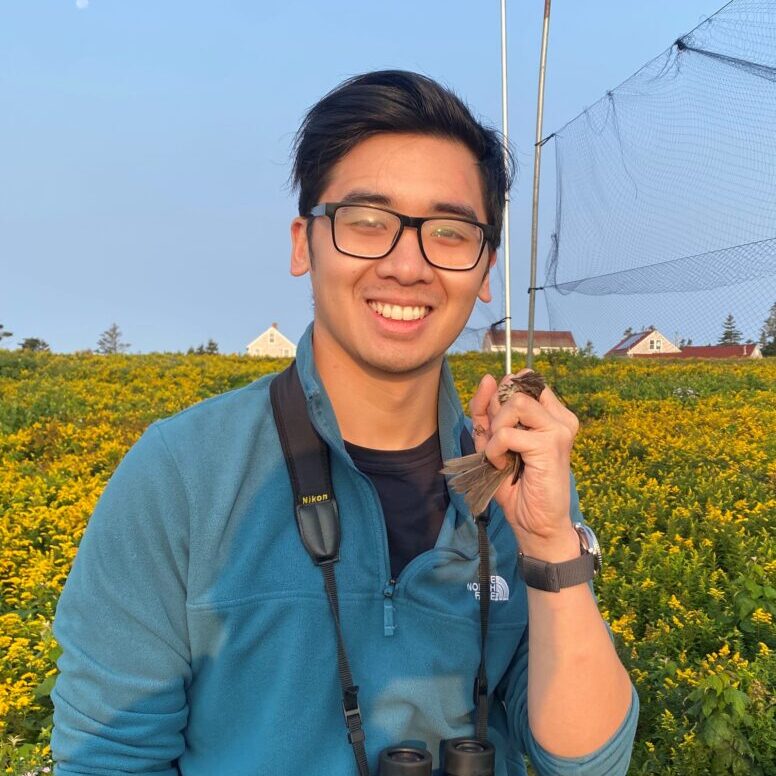 a person in a blue fleece sweater with black hair and glasses stands in a field with binoculars around his neck holding a small brown bird in his left hand