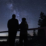 Silhouettes of two people standing in the dark looking at the night sky filled with stars.