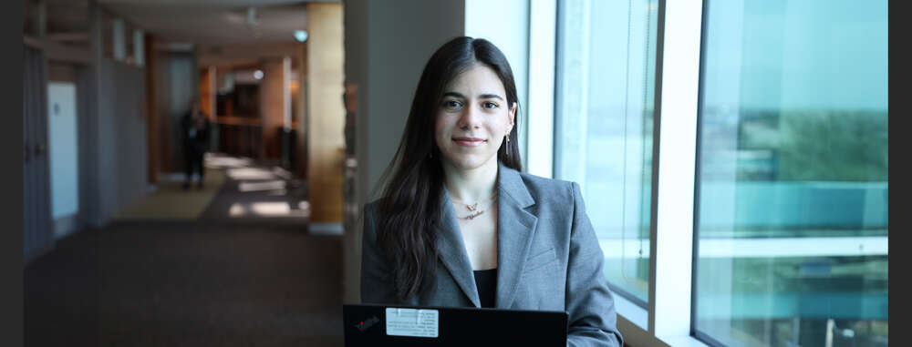 A co-op student wearing a suit stands in a hallway with an open laptop near a window