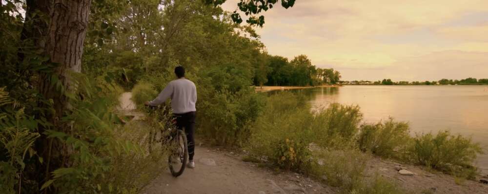 A person wearing a grey sweatshirt and black åsweatpants walks a black bicycle down a gravel path through greenery beside a lake as the sun sets in a pink sky.