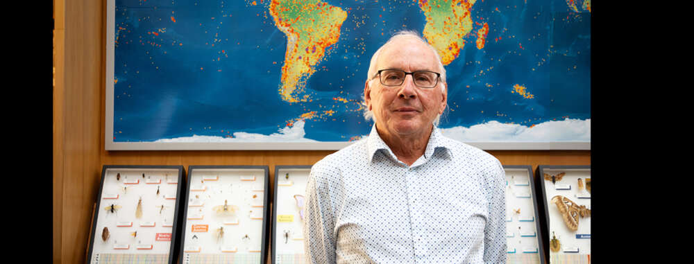 Dr. Paul Hebert poses for a portrait in the atrium of the Centre for Biodiversity Genomics at the University of Guelph with shadow boxes filled with insects on display behind him