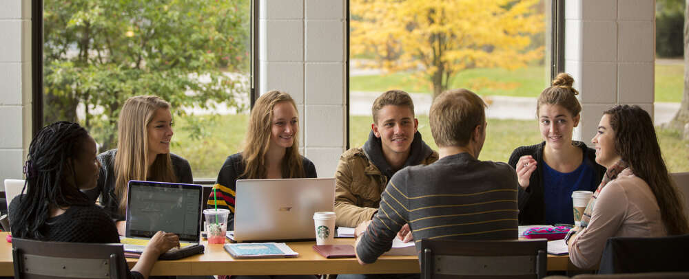A group of seven students sit facing one another at a table, some with their back to the camera, laughing and talking with laptops, notebooks and coffee cups on the table, in front of a window that overlooks the foliage on campus.
