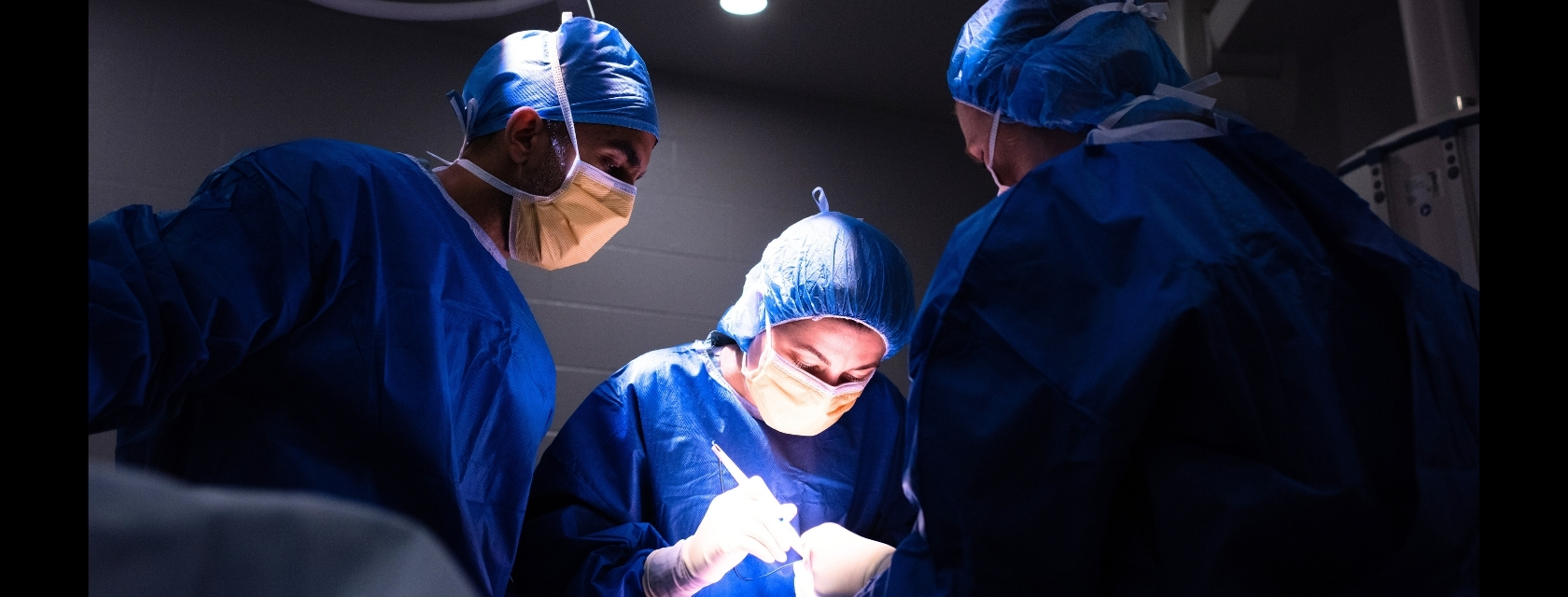 Surgeons in blue gowns perform operation under focused light.
