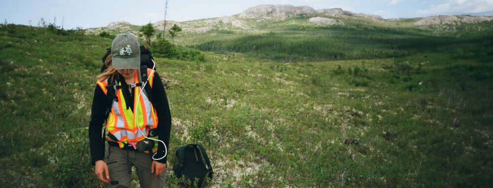 A person in a safety vest walks along a field of grass with mountains in the background