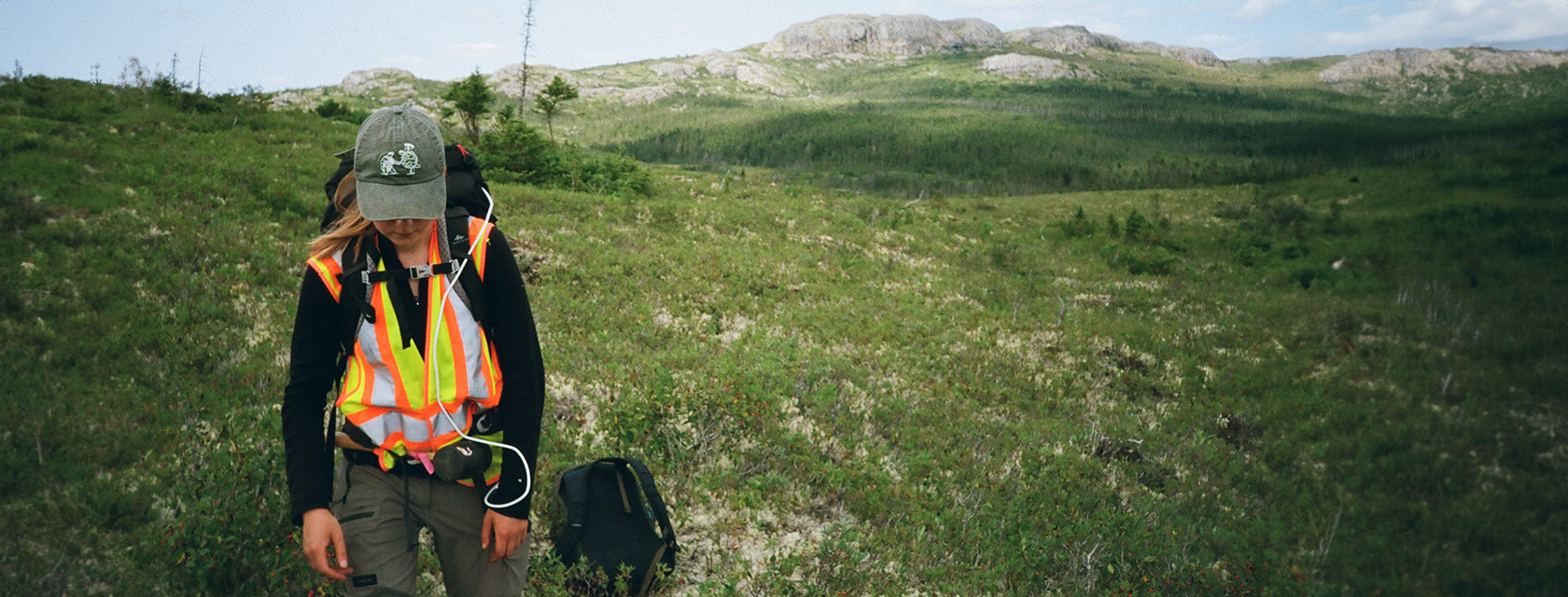 A person in a safety vest walks along a field of grass with mountains in the background