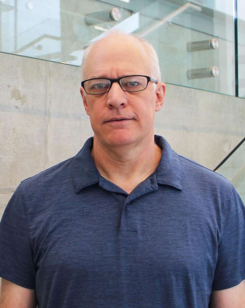 Person in navy shirt and glasses stands before glass stairwell.
