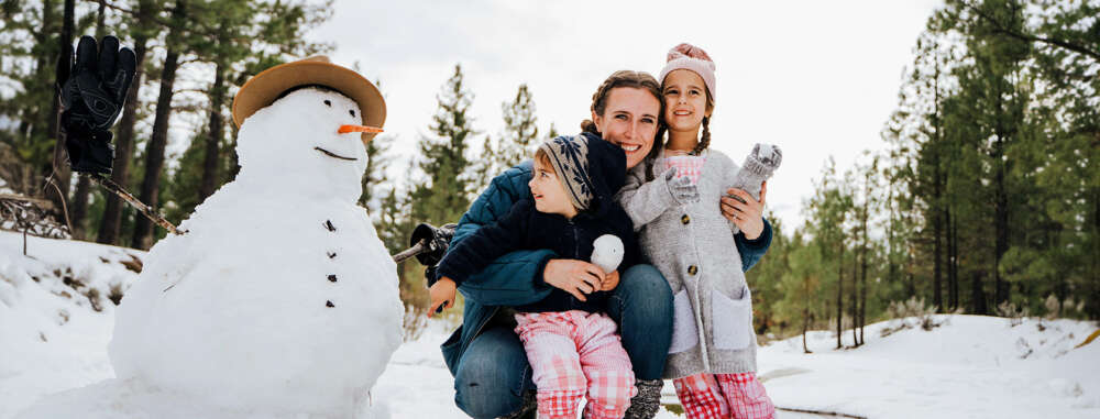 a woman and two kids smile posing beside a snowman on a snowy day near a forest