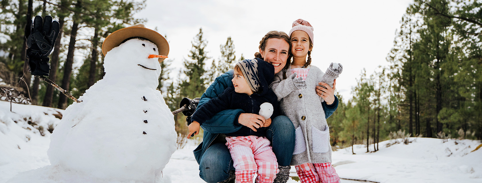 a woman and two kids smile posing beside a snowman on a snowy day near a forest