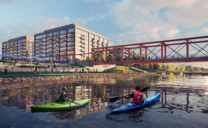 A pedestrian bridge carries people over a river next to a park, in the foreground kayakers paddle down a wide river.