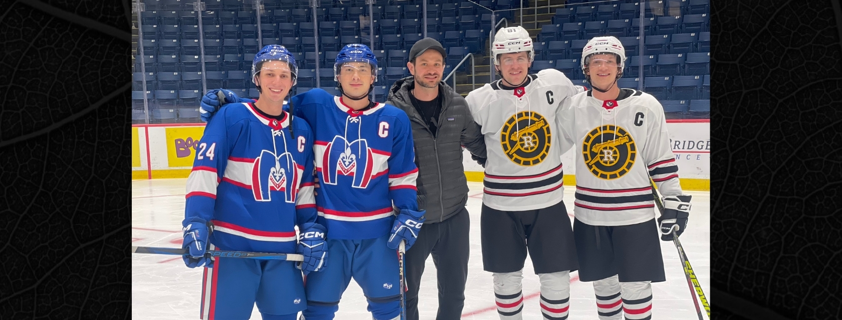 Two men stand on arena ice in hockey uniforms stand with a man in grey coat, black pants and black ball cap beside two men in white hockey uniforms smiling into the camera with arena stands behind them.