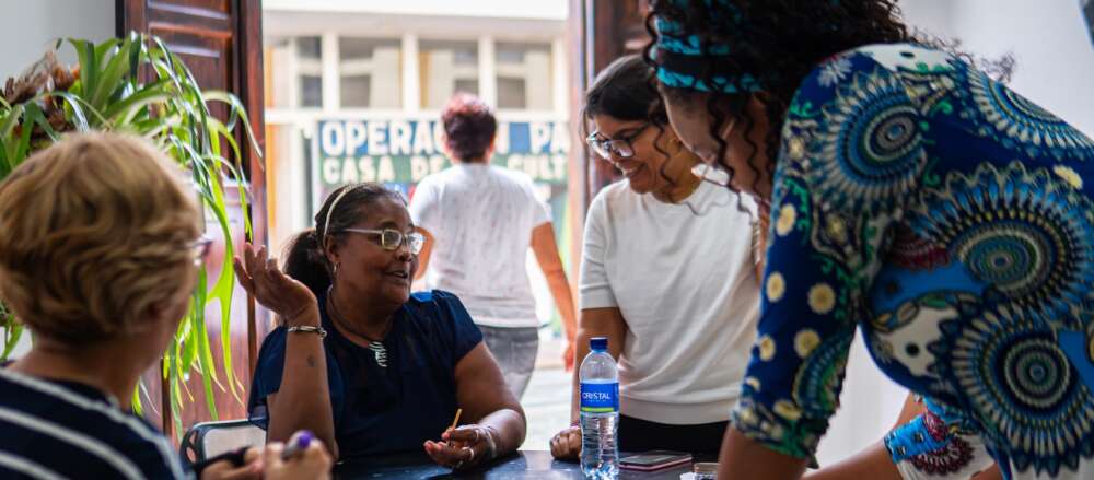A group of women dressed in blue and white gather around a table discussing as two are seated and two stand opposite.