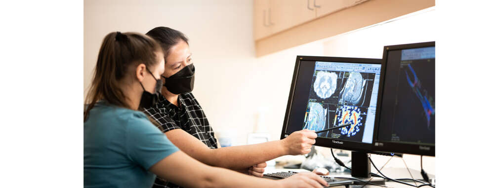 Two people wearing black face masks look at two computer screens showing medical imaging