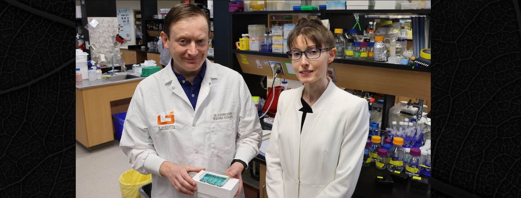 Two researchers in white lab coats over dark-coloured clothing stand facing the camera smiling and one holds a blood sample.
