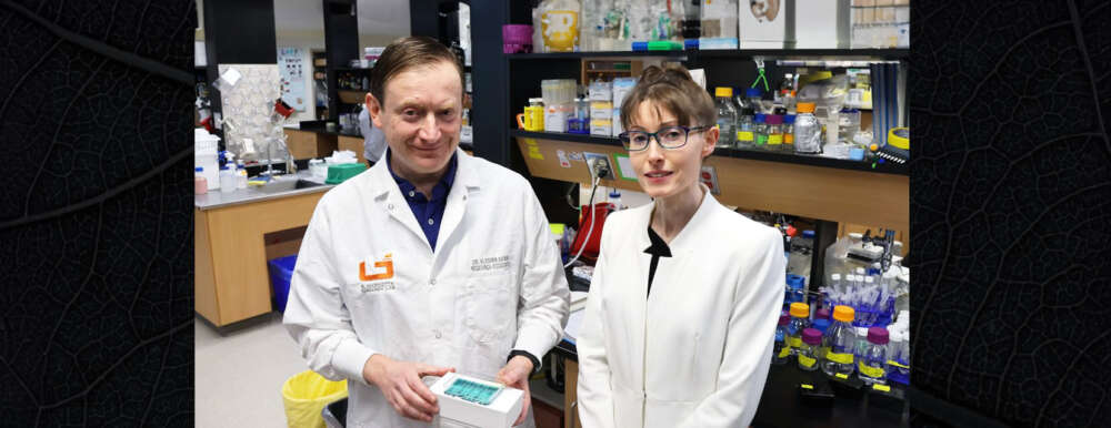 Two researchers in white lab coats face the camera smiling and one holds a blood analysis device.