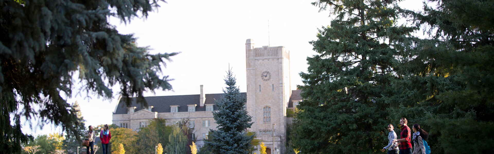 Students walk across green campus space known as Johnston Green in front of gray building Johnston Hall in the summer.