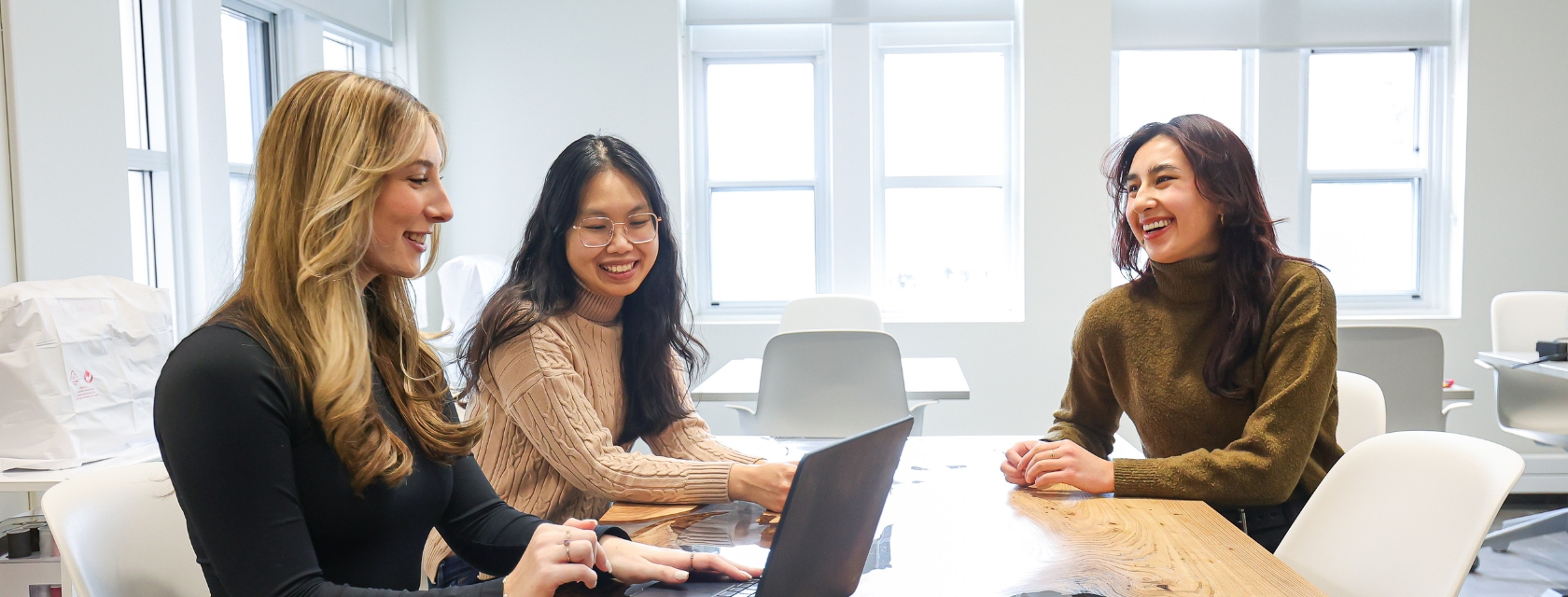 Three students sit in a brightly lit office discussing something over a laptop