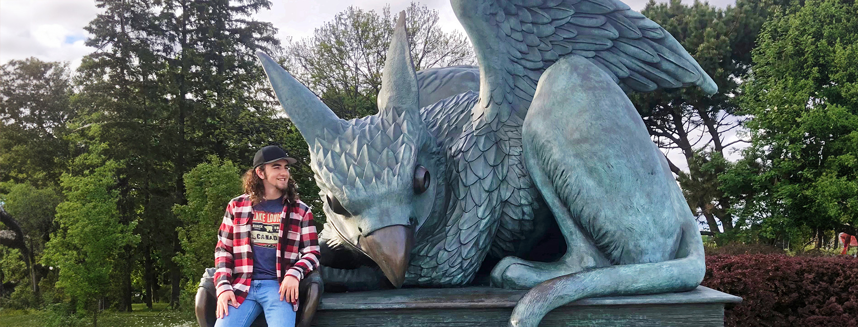 person in plaid shirt sits beside a large statue of a gryphon with wings and beak